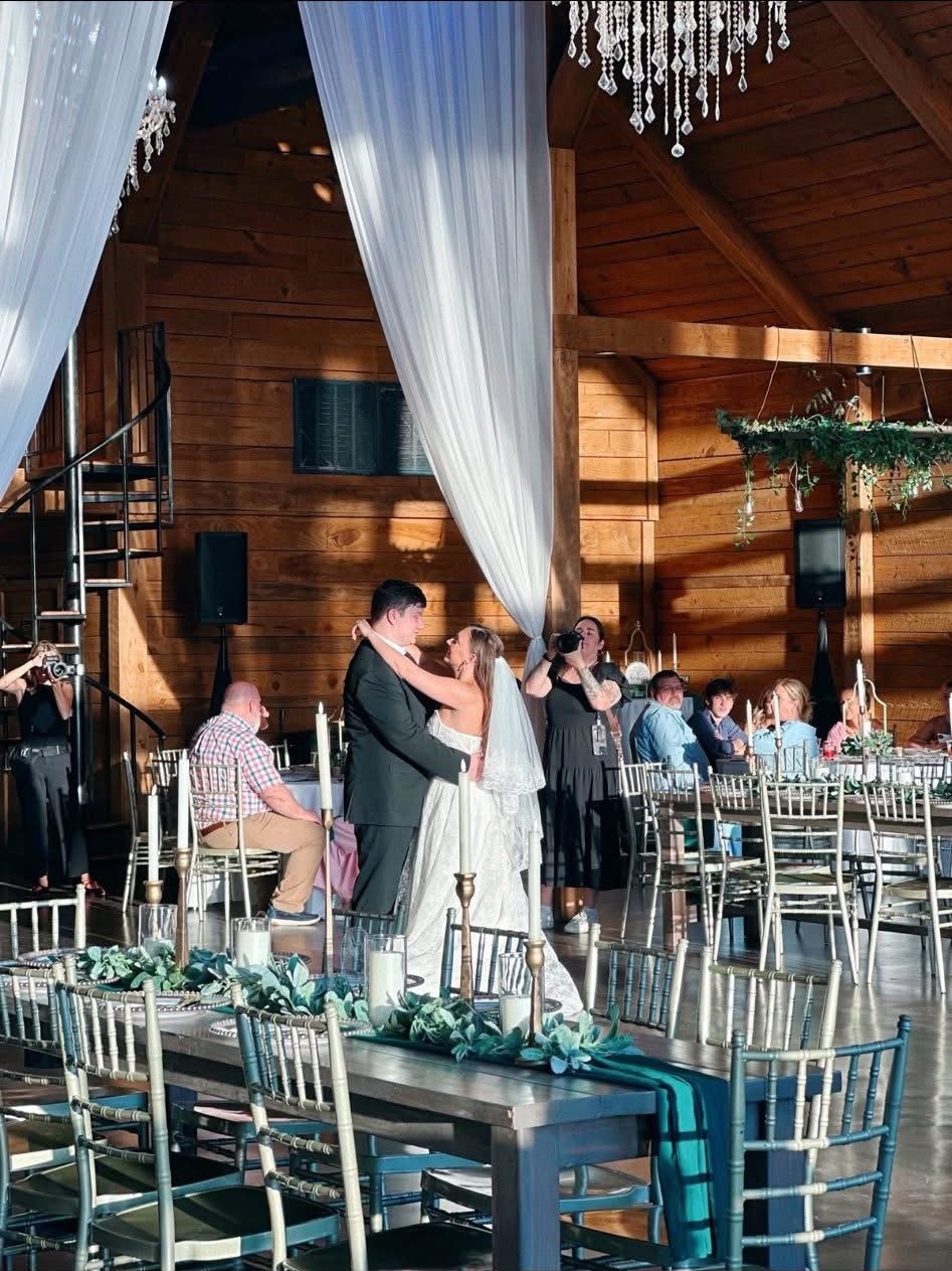 Bride and groom's first dance under draped fabric in a rustic barn venue, with a Rivera Event Sound speaker on a stand in the background
